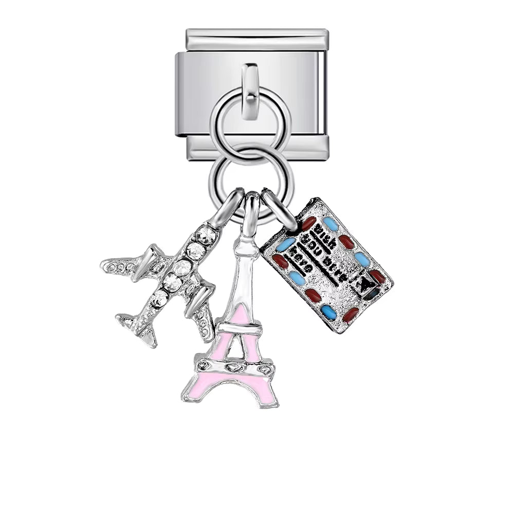 Silver charm with multiple charms including a pink Eiffel Tower, a suitcase, and a heart with a key on a white background.